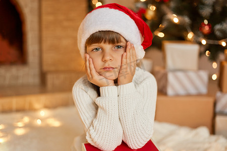 等待圣诞节的无聊小女孩，坐在喜庆的客厅里，双手放在脸颊上，穿着白色毛衣和红色圣诞帽，用悲伤的表情看着镜头。