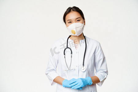 戴着呼吸器、医用口罩和橡胶手套的亚洲女医生面带微笑，身穿白色背景的诊所制服，随时准备提供帮助。 
