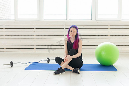 留着紫色头发的年轻混血女嬉皮士坐在杠铃旁边的运动垫上，在健身房里拿着健身球。