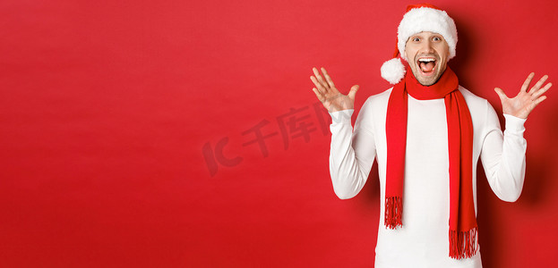 快乐而惊奇的帅哥的画像，庆祝新年，祝圣诞快乐，戴着圣诞帽和围巾，讲述重大新闻，站在红色背景下