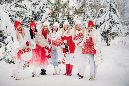 一大群女孩手里拿着圣诞礼物站在冬天的森林里。穿着红白相间衣服的女孩在白雪皑皑的森林里拿着圣诞礼物