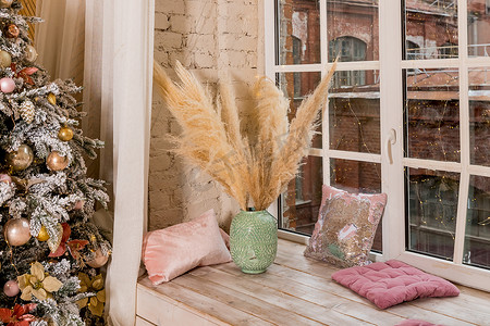 圣诞节花瓶摄影照片_在白色窗台上的绿色花瓶中的潘帕斯草花束在家里的花环的窗边。圣诞节组成。