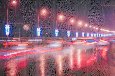 在被雨水覆盖的窗户后面的夜城里，移动的汽车和灯笼的模糊灯光反射在潮湿的沥青上。