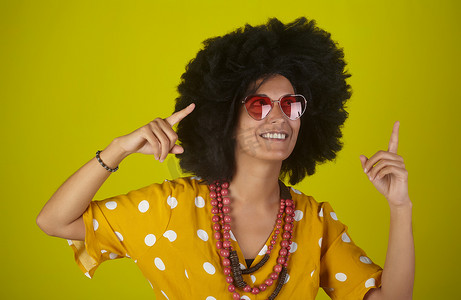 美丽而微笑的女人，留着卷曲的非洲发型，戴着心形太阳镜，用黄色背景上的手指向右看和指示方向