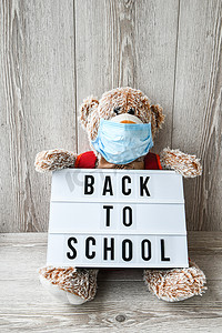 泰迪熊玩偶戴着面罩背包，手拿灯箱，灯箱上写着木制背景的“BACK TO SCHOOL”文字，复制空间，2020 年隔离。新常态