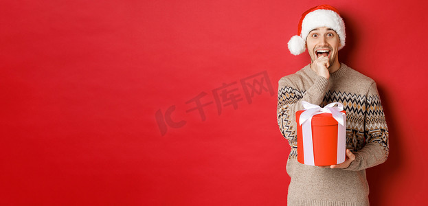 戴着圣诞帽和圣诞毛衣的帅哥兴奋地打开圣诞礼物，看上去很惊讶，一只手拿着礼物，站在红色背景上