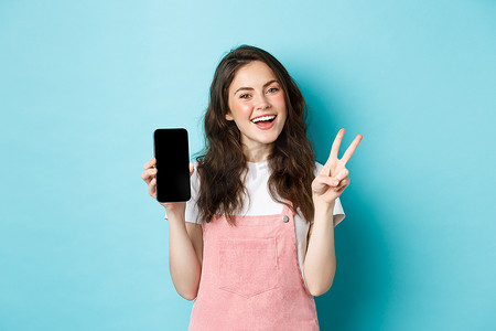 app展示摄影照片_可爱的年轻女性微笑着，用空的智能手机屏幕展示 v 形标志，展示应用程序或移动商店，站在蓝色背景下