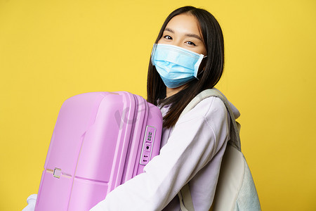戴着医用面罩、拿着粉色可爱手提箱、去度假、在流行病期间旅行、黄色背景的快乐亚洲女游客的特写