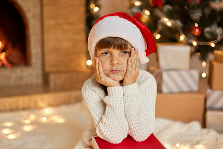 表情装饰摄影照片_戴着圣诞帽和白色毛衣的小女孩坐在圣诞树旁，手里拿着成堆的礼物盒，表情沉思，看着镜头，双手放在下巴下。