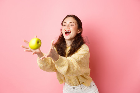 快乐可爱的女孩抓起一个苹果笑着，是时候享用充满水果的健康午餐，站在粉红色背景下