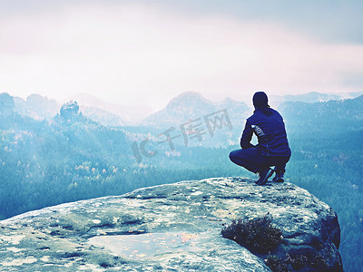 男士风摄影照片_黑衣游客坐在悬崖边缘，望着迷雾笼罩的山谷