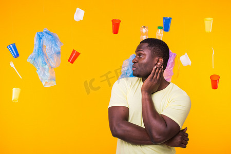 世界环境日、塑料回收问题和环境灾难概念 — 黄色背景下看着垃圾的非洲裔美国人感到惊讶。
