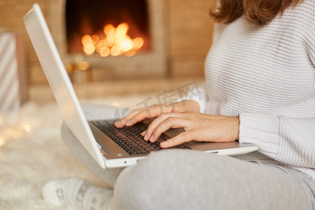 不知火摄影照片_匿名女性在她的笔记本电脑上打字，她在温暖的火炉旁放松，不知名的女性在网上工作，双腿交叉坐在地板上，膝盖上拿着笔记本。