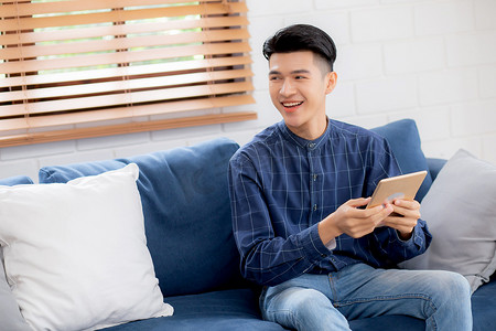 阅读交流摄影照片_年轻有魅力的亚洲男人在家里的沙发上使用浏览平板电脑休息，快乐的男性坐在沙发上放松阅读家里的数字小工具、交流和生活方式概念。