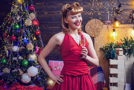微笑的 ponup 女人穿着红色晚礼服，在家里的圣诞树背景上拿着一杯起泡酒。