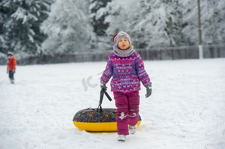 紫色衣服摄影照片_一个冬天穿着紫色衣服和充气圈的小女孩在白雪覆盖的森林里走在街上