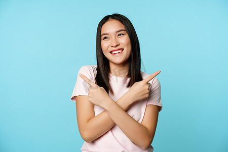 微笑的亚洲女孩侧指，展示两种选择，笑着，在变体之间挑选，蓝色背景