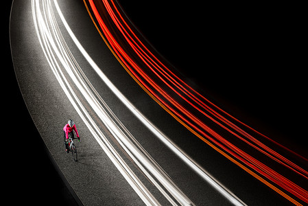 女骑自行车的人在夜间道路上骑自行车，有汽车灯迹。