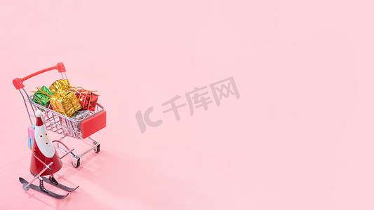 圣诞购物概念、带圣诞老人玩具的迷你红色购物车手推车和淡粉色背景中突显的礼盒、空白复制空间、特写