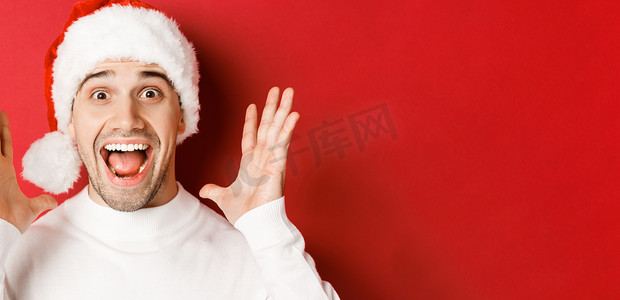 戴着圣诞帽的快乐年轻人的特写，发布重大的圣诞公告，微笑着惊讶，站在红色背景上