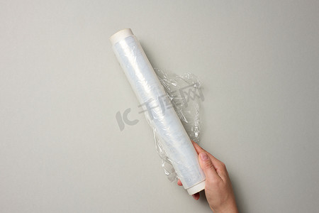 透明灰色背景摄影照片_两只女手拿着一卷透明保鲜膜，用于包装产品，灰色背景
