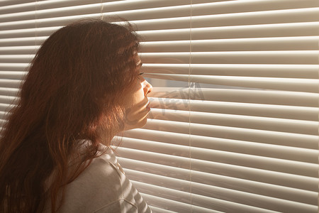 美丽的年轻女子的后视图，长发，透过百叶窗上的洞，望向窗外。