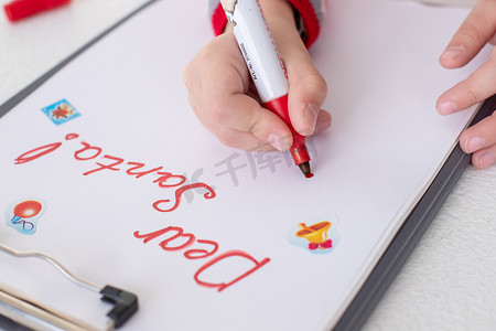新年目标摄影照片_亲爱的圣诞老人信，圣诞卡。女孩穿着红色毛衣，拿着毡笔，在白色背景的白纸上写字。关于礼物的童年梦想。圣诞节和新年概念。寒假
