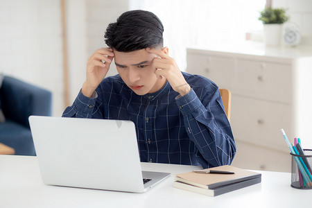 年轻的亚洲商人在笔记本电脑上工作时头痛，最后期限在家里的办公桌上，失败的商人筋疲力尽、疲倦、压力和担心、沮丧和失败。