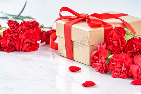 丝带感恩背景摄影照片_母亲节问候设计理念 — 美丽盛开的红色康乃馨，白色大理石桌背景上带丝带的礼盒，特写，复制空间。