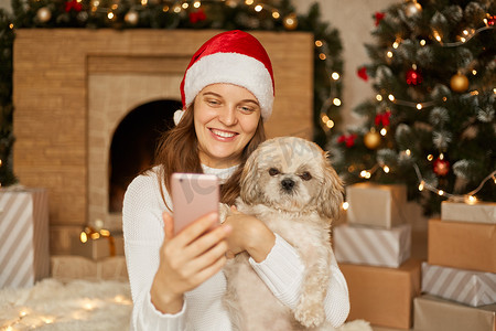 女孩坐在带壁炉和圣诞树的节日房间里与漂亮的小狗自拍，女孩戴着圣诞帽和休闲套头衫，带着迷人的微笑看着手机屏幕。