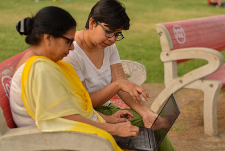 在印度新德里的一个公园里，年轻的印度女孩在坐在红色长椅上的笔记本电脑上帮助一位印度老妇人的侧视图。