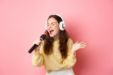年轻女子唱卡拉 OK、戴着耳机和表演歌曲、手持麦克风、站在粉红色背景下的肖像