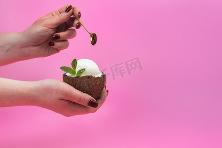 手拿薄荷叶摄影照片_香草冰淇淋球放在新鲜椰子的一半里，手里拿着薄荷叶装饰，粉色背景上放着勺子