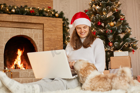 迷人的女孩带着笔记本电脑和狗在圣诞节的客厅里，女士看着她的宠物微笑，假期在家工作的自由职业者，穿着白色休闲白色套头衫和红色帽子的女孩。