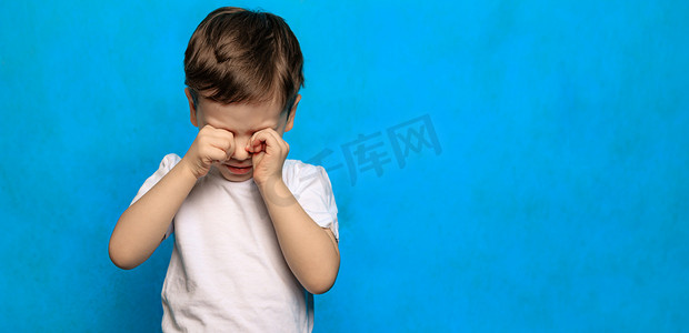 哭眼睛摄影照片_蓝色背景上的一个男孩揉着眼睛。