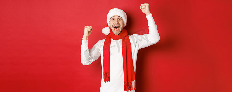 戴着圣诞帽和围巾的快乐白人男子的形象，欢呼雀跃，举起双手，庆祝胜利或胜利，战胜红色背景