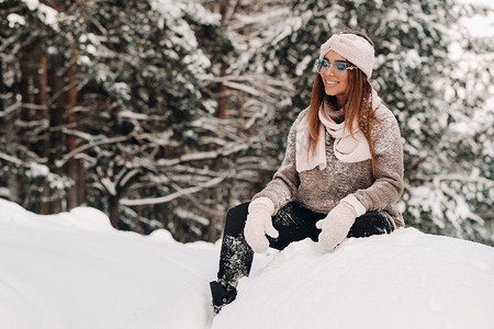 毛线毛衣摄影照片_冬天，一个穿着毛衣和眼镜的女孩坐在森林里冰雪覆盖的背景上