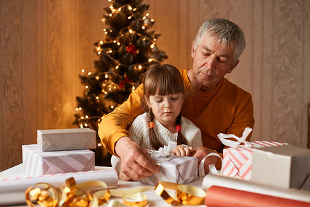 室内照片中，一位身穿橙色毛衣的老人和他的孙女跪在桌旁，为圣诞节假期打包礼物盒。