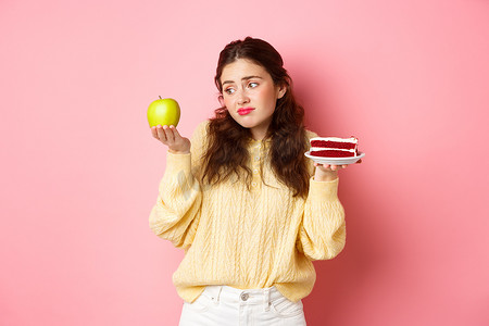 女孩美味表情摄影照片_年轻女子面带悲伤和失望的表情看着青苹果，同时拿着一块美味的蛋糕，站在粉红色的背景下，想吃甜点
