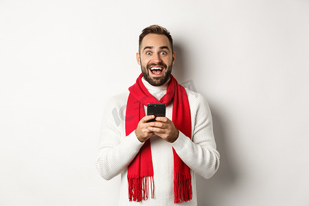 促销冬季摄影照片_男人在互联网上阅读令人惊叹的促销优惠，拿着智能手机，看上去很惊讶，站在白色背景下的冬季毛衣