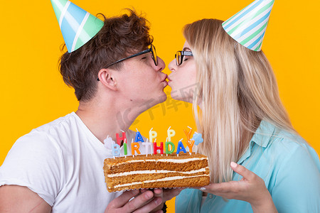 快乐的一对迷人的男人和戴着纸帽的可爱的女孩做出愚蠢的表情，手里拿着蛋糕，黄色背景上刻着生日铭文。