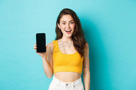 身穿黄色短上衣的兴奋的年轻女性展示空白的智能手机屏幕，展示您展示的广告，站在蓝色背景上