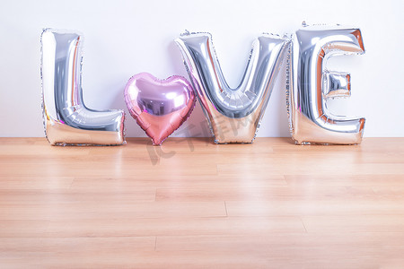 母亲字摄影照片_情人节、母亲节设计理念 — 浅色木地板和白墙背景上带有爱字形状的漂亮气球，特写