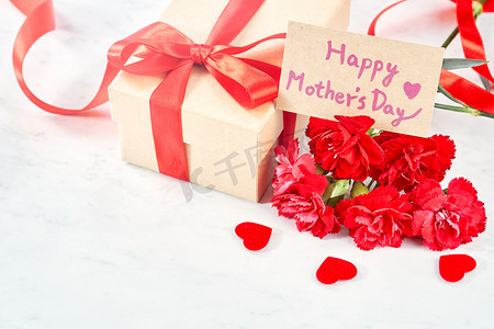 丝带感恩背景摄影照片_母亲节问候设计理念 — 美丽盛开的红色康乃馨，白色大理石桌背景上带丝带的礼盒，特写，复制空间。