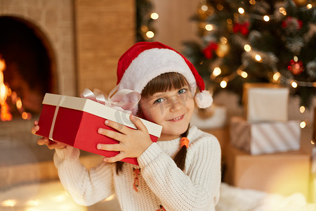 戴着圣诞老人红帽子的有趣的孩子手里拿着圣诞礼物，直视相机，试图猜测礼物盒里有什么，坐在客厅靠近壁炉和圣诞树的地方。