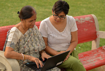 在印度新德里的一个公园里，年轻的印度女孩在坐在红色长椅上的笔记本电脑上帮助一位印度老妇人的侧视图。