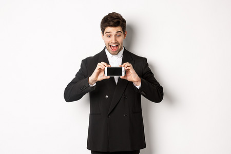 兴奋的帅哥展示智能手机屏幕的肖像，看着广告展示，穿着黑色西装站在白色背景上，看起来很惊讶