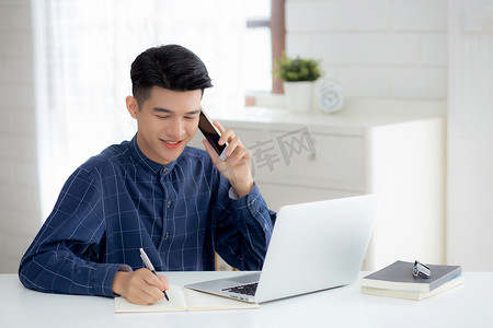 年轻的亚洲男子通过笔记本电脑在家打电话和工作，自由男性在笔记本上写作并在智能手机上讲话，呆在家里，家庭生活，商业和沟通理念。