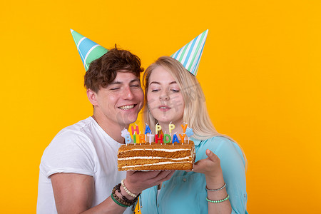 黄色字样摄影照片_一对年轻迷人的男人和戴着纸帽的可爱女孩做傻脸，手里拿着蛋糕，黄色背景上刻着生日字样。