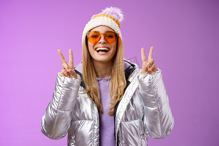 戴着墨镜、银色时尚夹克、冬帽、友好、幸运、乐观、美丽的金发美女展示和平胜利姿态，享受雪地假期，到酒店外享受滑雪，紫色背景